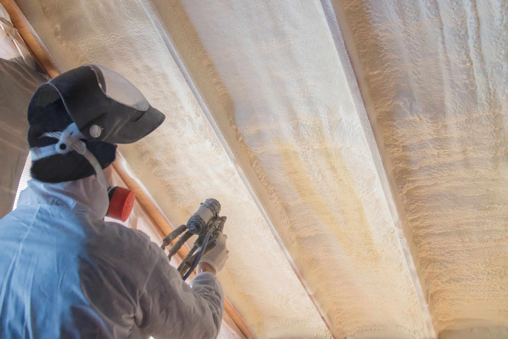 Ouvrier équipé d'un masque de protection et combinaison appliquant de l'isolant thermique par projection sur chevrons de toiture en construction intérieure