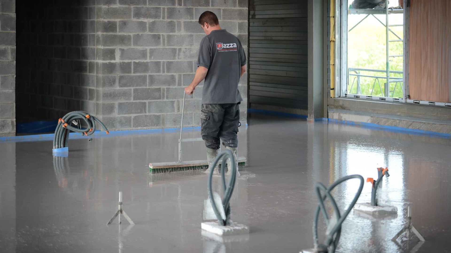 Technicien en tenue de travail versant une chape fluide ciment gris clair depuis un tuyau de pompage sur une surface isolante préparée, avec plots de nivellement métalliques visibles et surface réfléchissante du mortier frais en phase d'autonivelage dans un intérieur de chantier à finir.